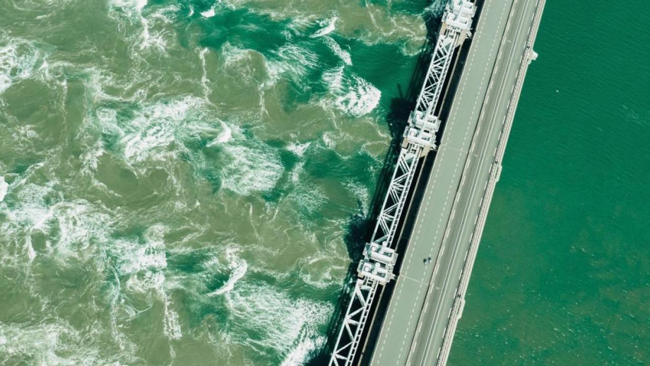 Aerial view of a large dam with green water on one side and cloudy green turbulent water on the other.