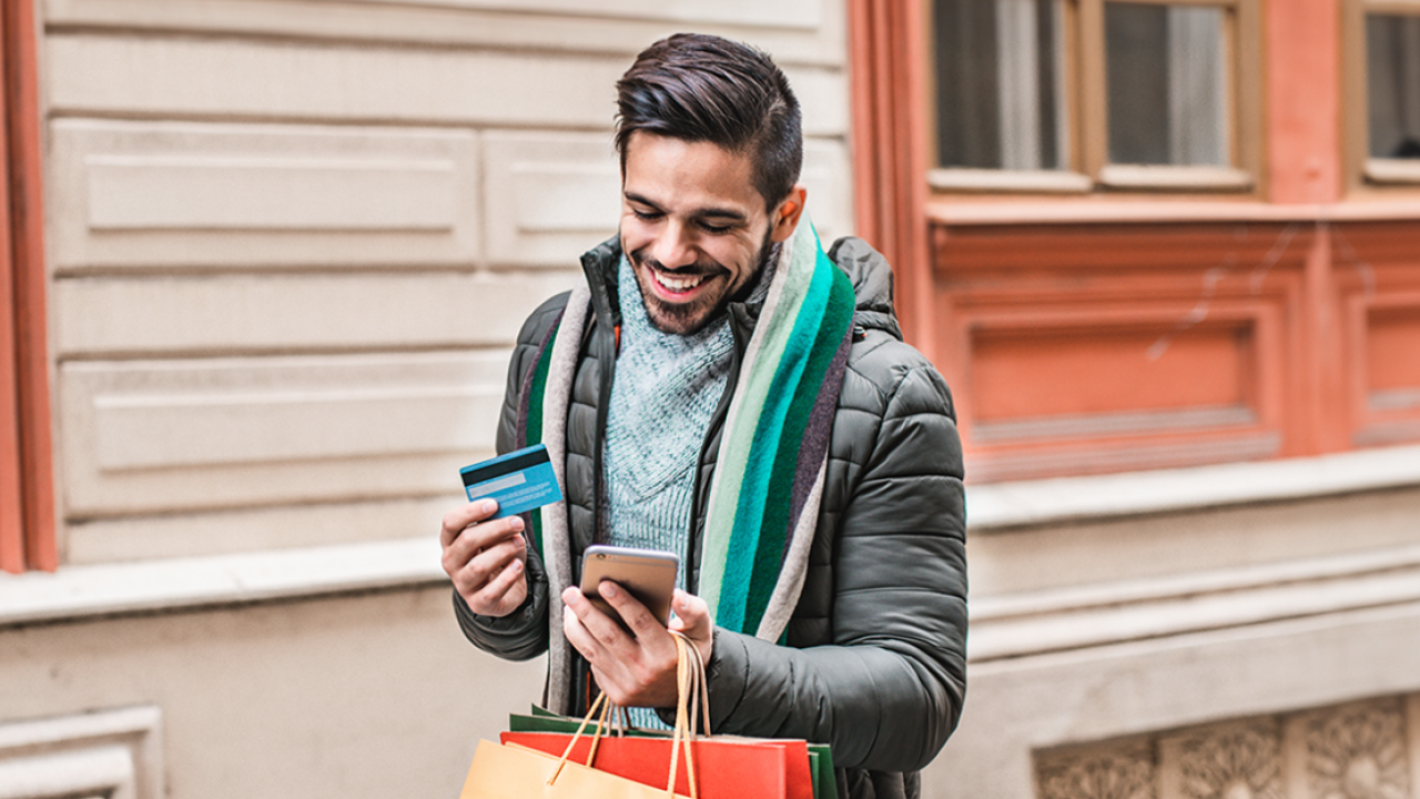 A smiling person outside holding a credit card and cellphone and shopping bags.