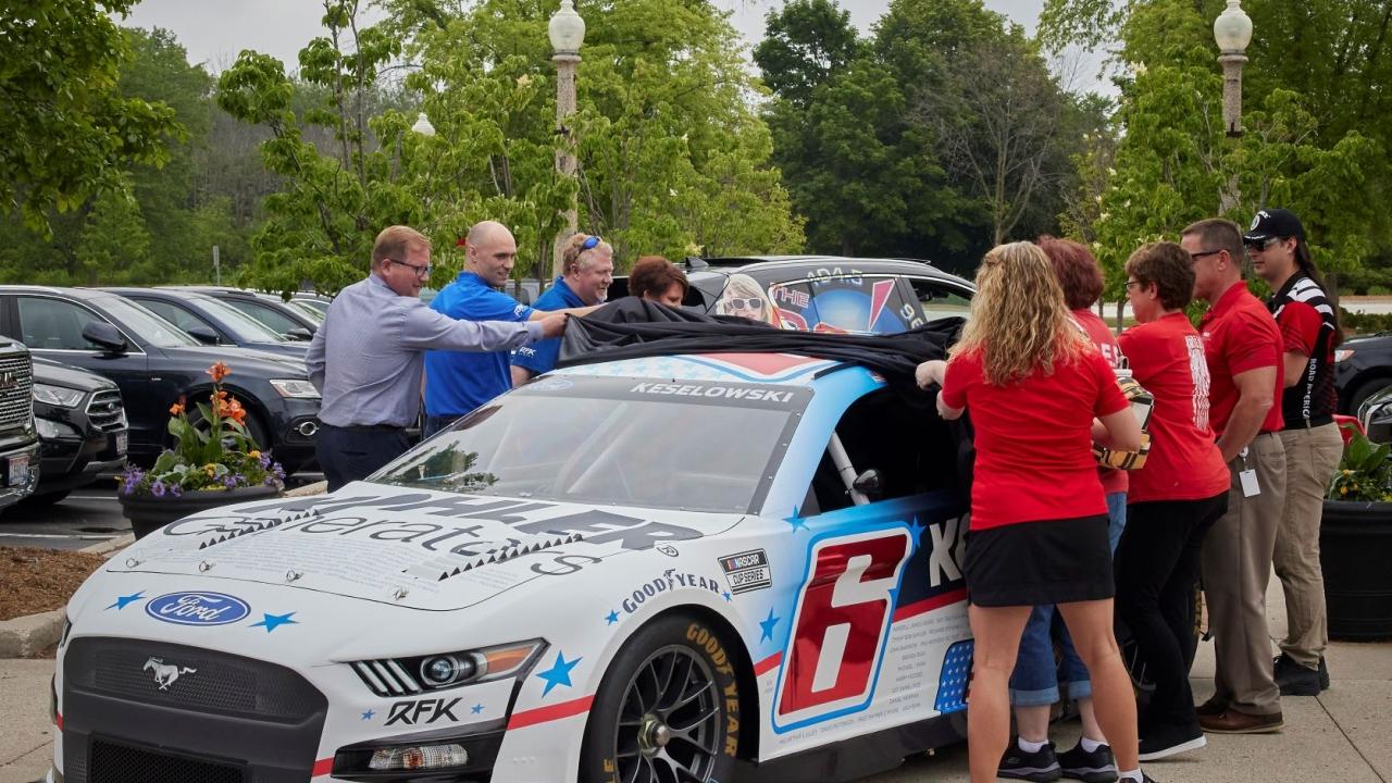 a group of people stand outside and take a cover off a race car. "6" on the side