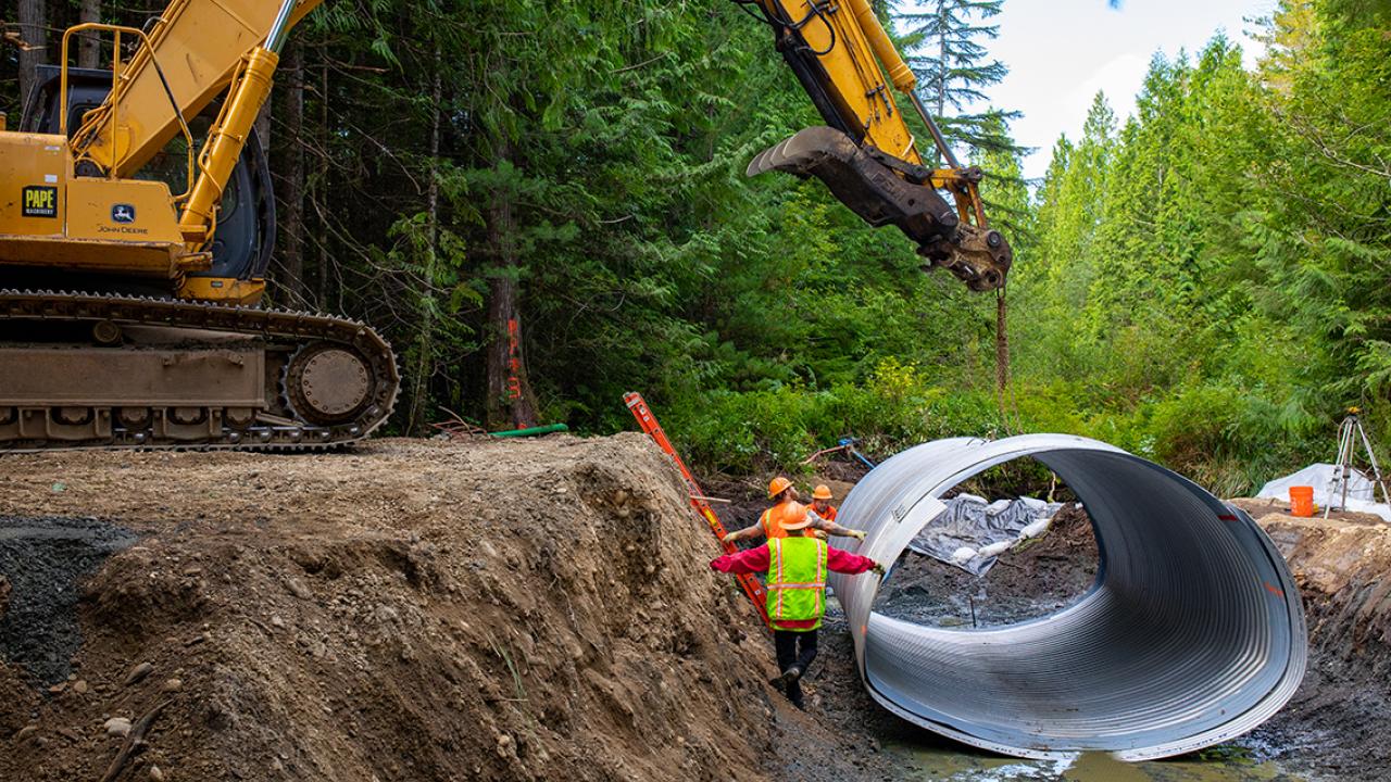 A large machine lifting a metal culvert as workers in a ditch position it.
