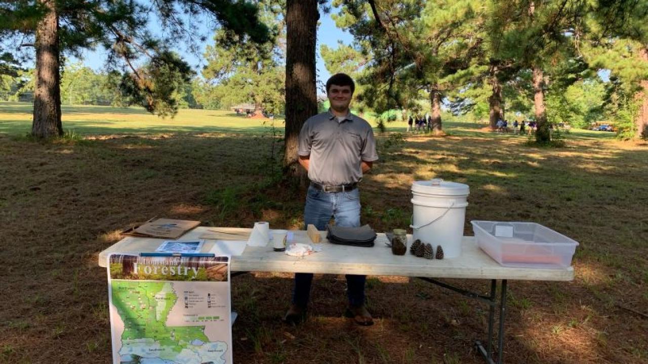 A person standing in a park behind a table with presentation materials and a map of Louisiana.