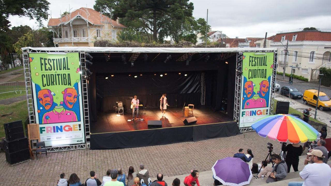 Two people performing on an outdoor stage, an audience in cement bleachers.