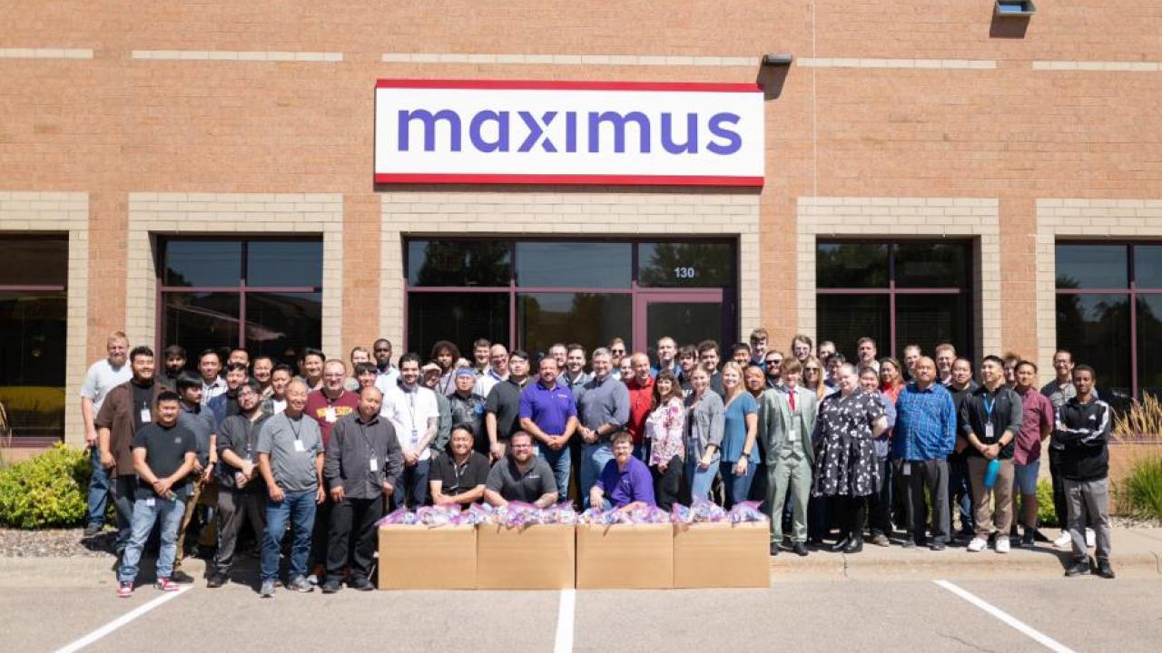 A large group posed outside a Maximus building, packed boxes in front of them.