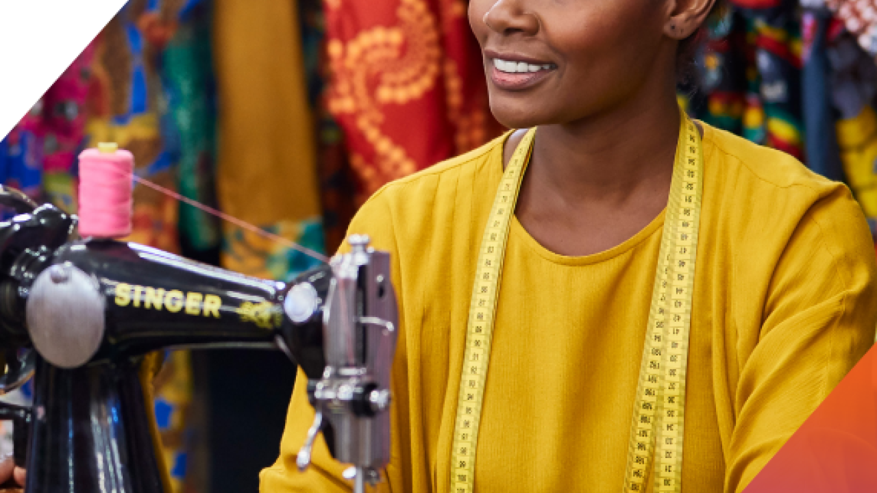 A person sitting behind a sewing machine. Bright colored fabrics behind them.