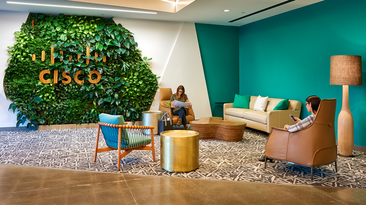 people sitting in a living room setting, a wall of greenery behind them with Cisco logo on it