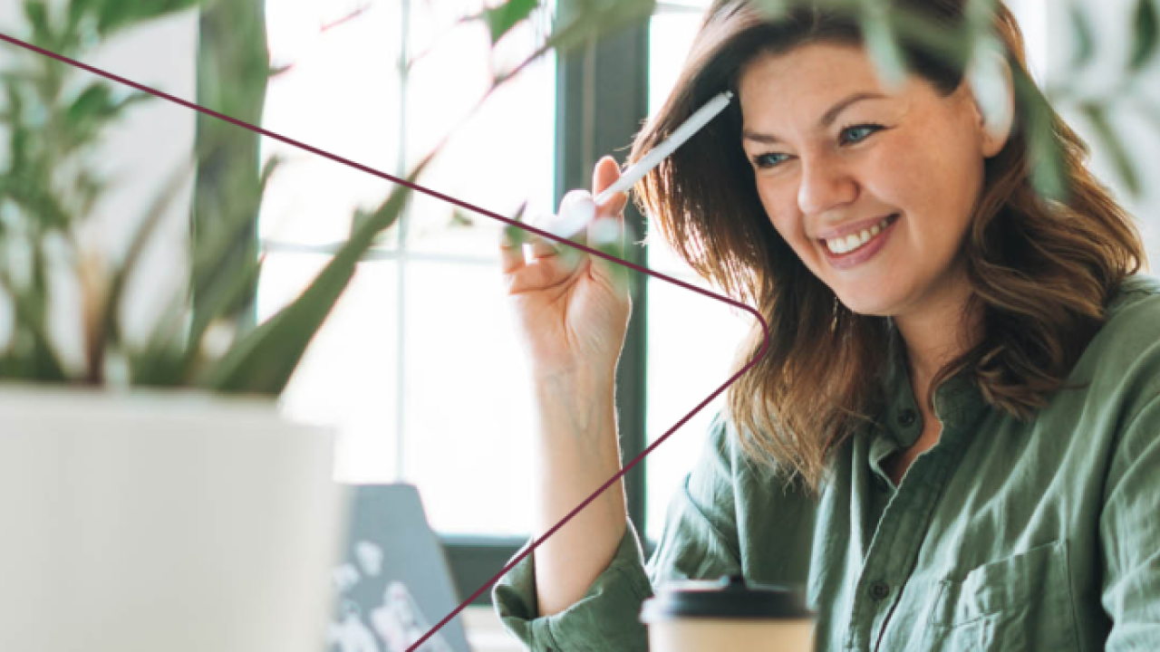 A smiling person looking at a laptop, a pen by their head.
