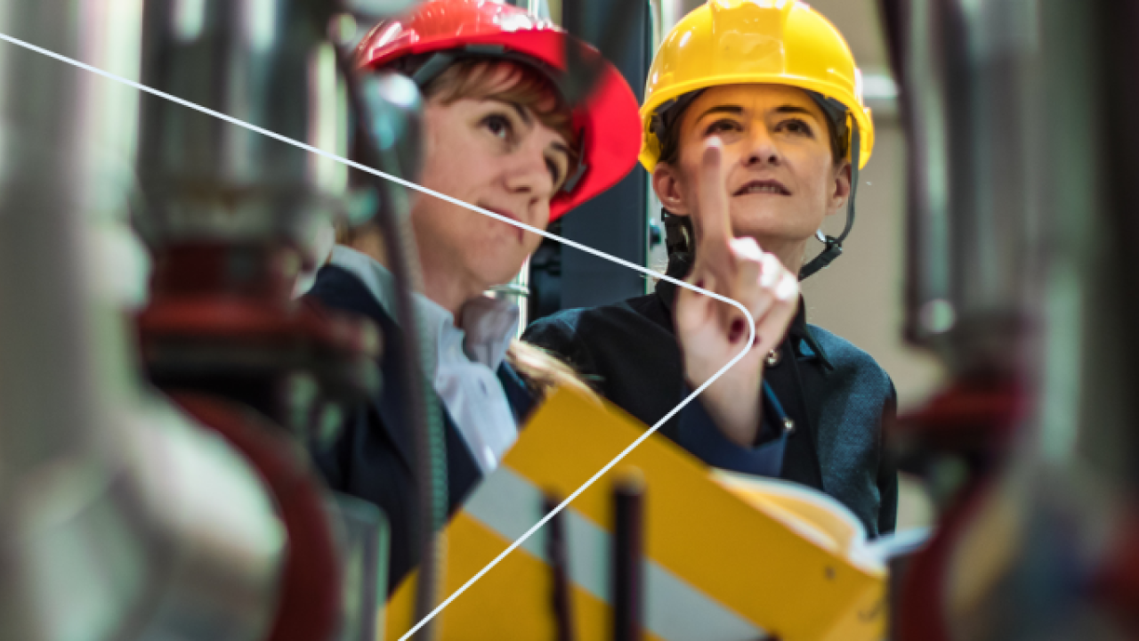 Two people in hard hats in the midst of industrial equipment and piping.