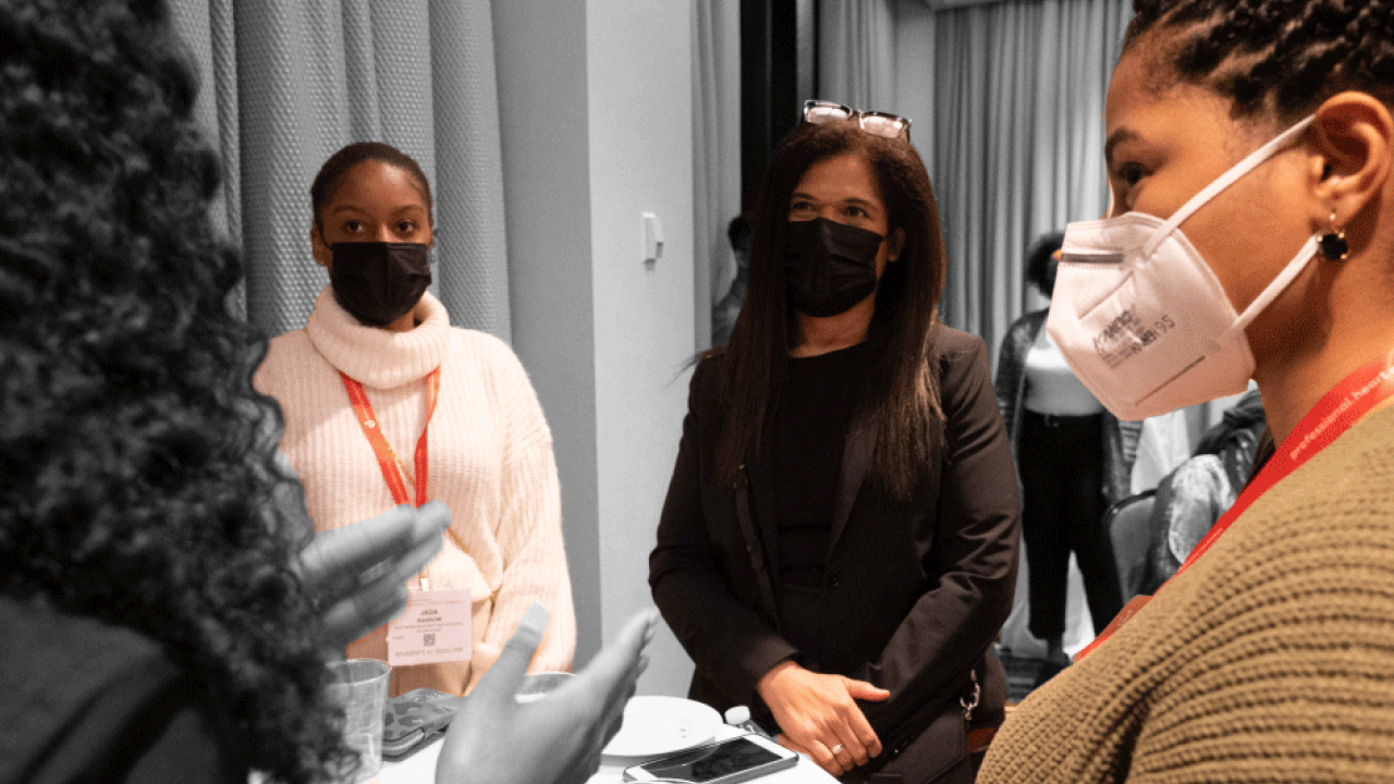 a group of people stand around a small round table, all wearing protective masks