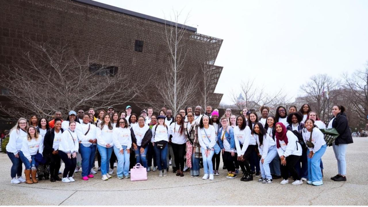 Over 50 Bath & Body Works associates are smiling pictured together in matching shirts in front of the National Museum of African American History and Culture. The museum building is in the background.