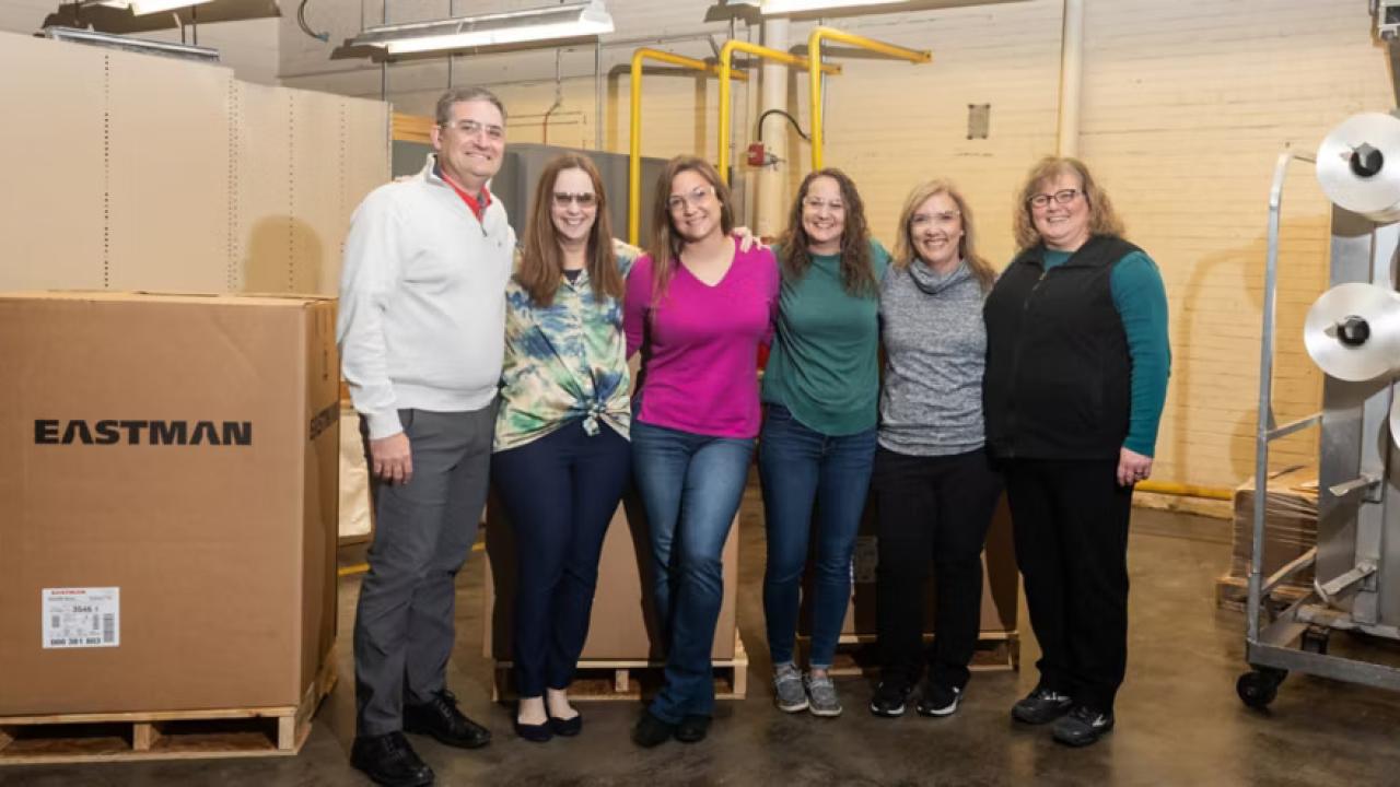 Six people posed with arms around each other. Large spools of thread to the side and an Eastman Box on the other side.