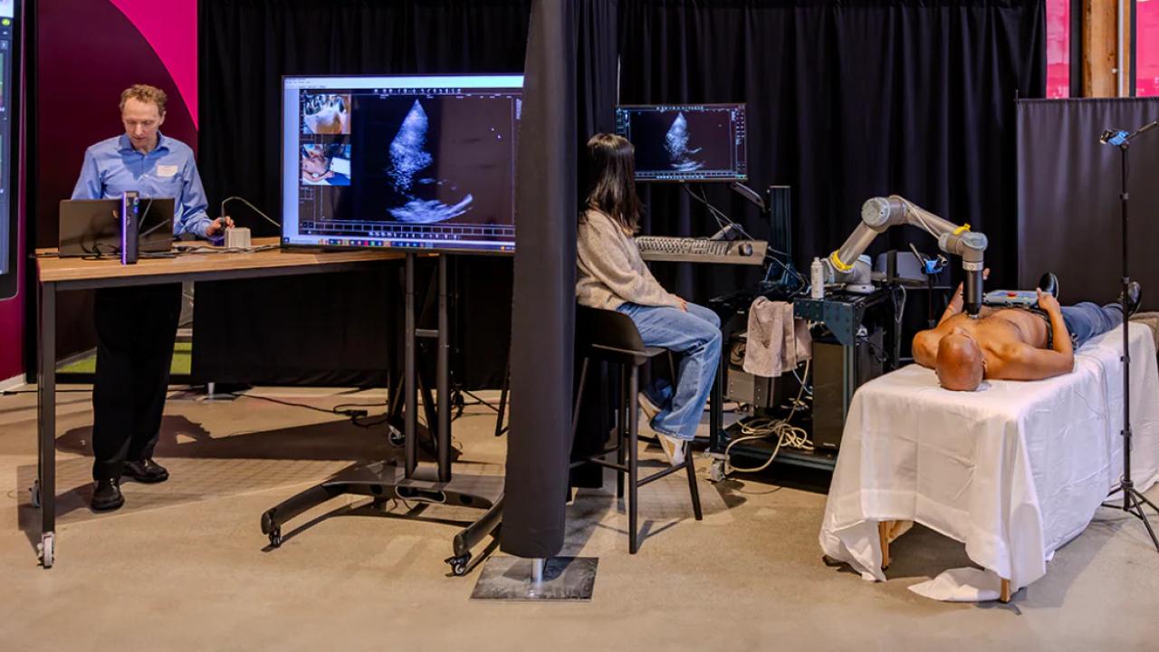 A person on a table with another sitting nearby. A medical imaging device on their chest. Another person behind a curtain viewing the images.
