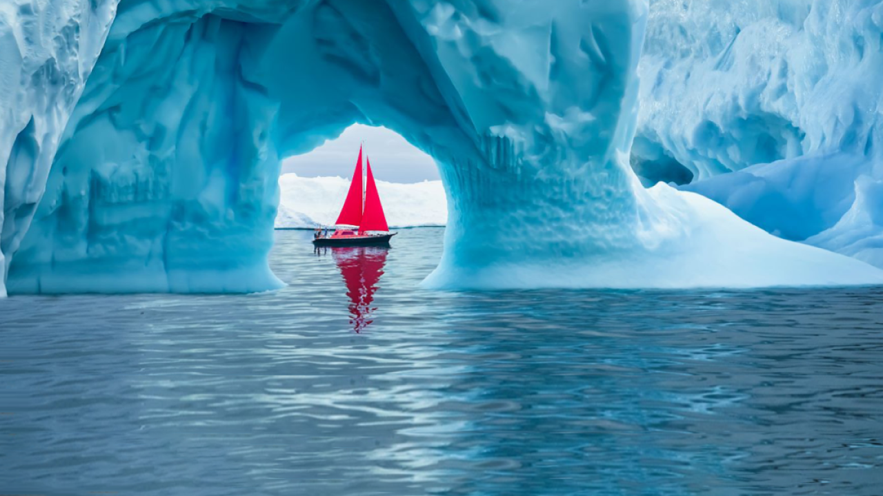 A boat with red sails framed in a small opening in an iceberg on a body of water.