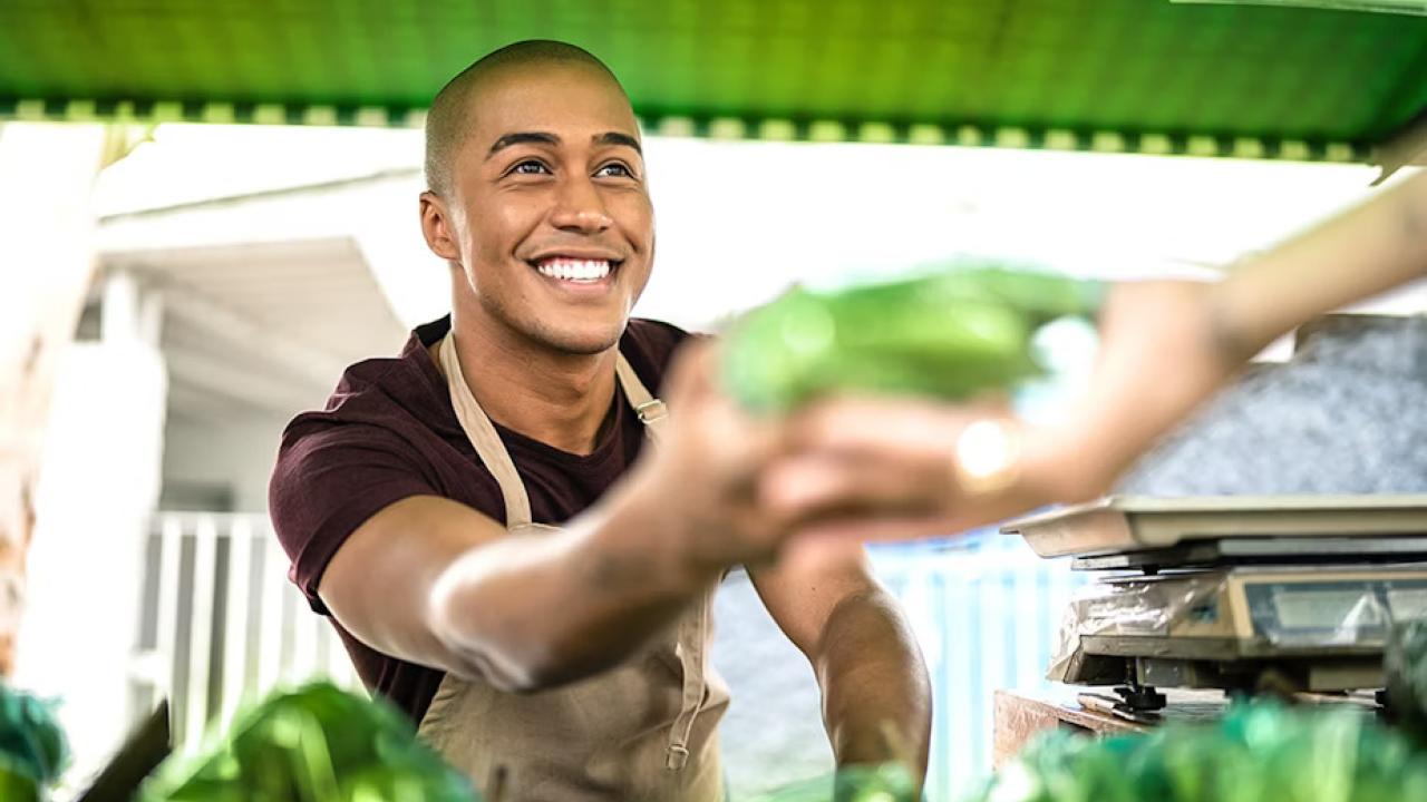 A smiling person in an apron handing something green to another.