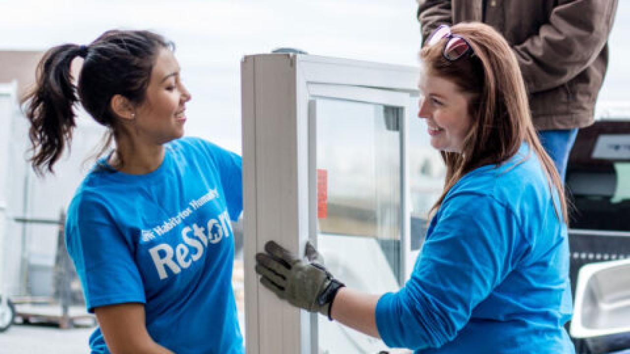 Two people wearing blue tops, working together to carry a large item