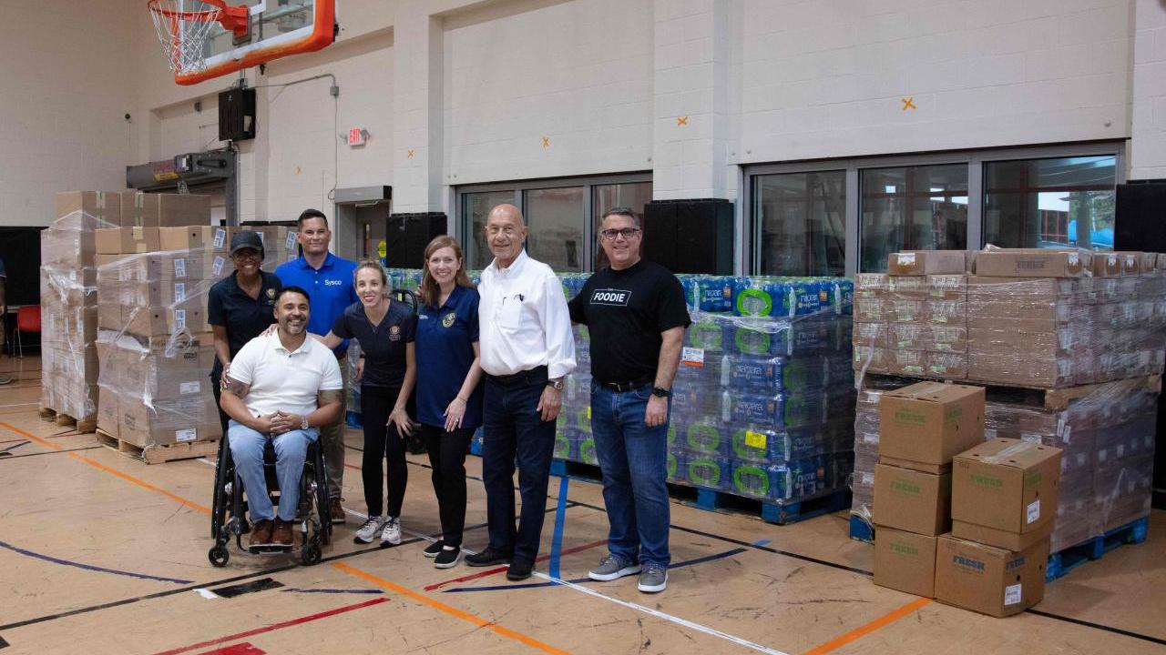 People posed in front of pallets of boxes of food, water and supplies in a gymnasium.