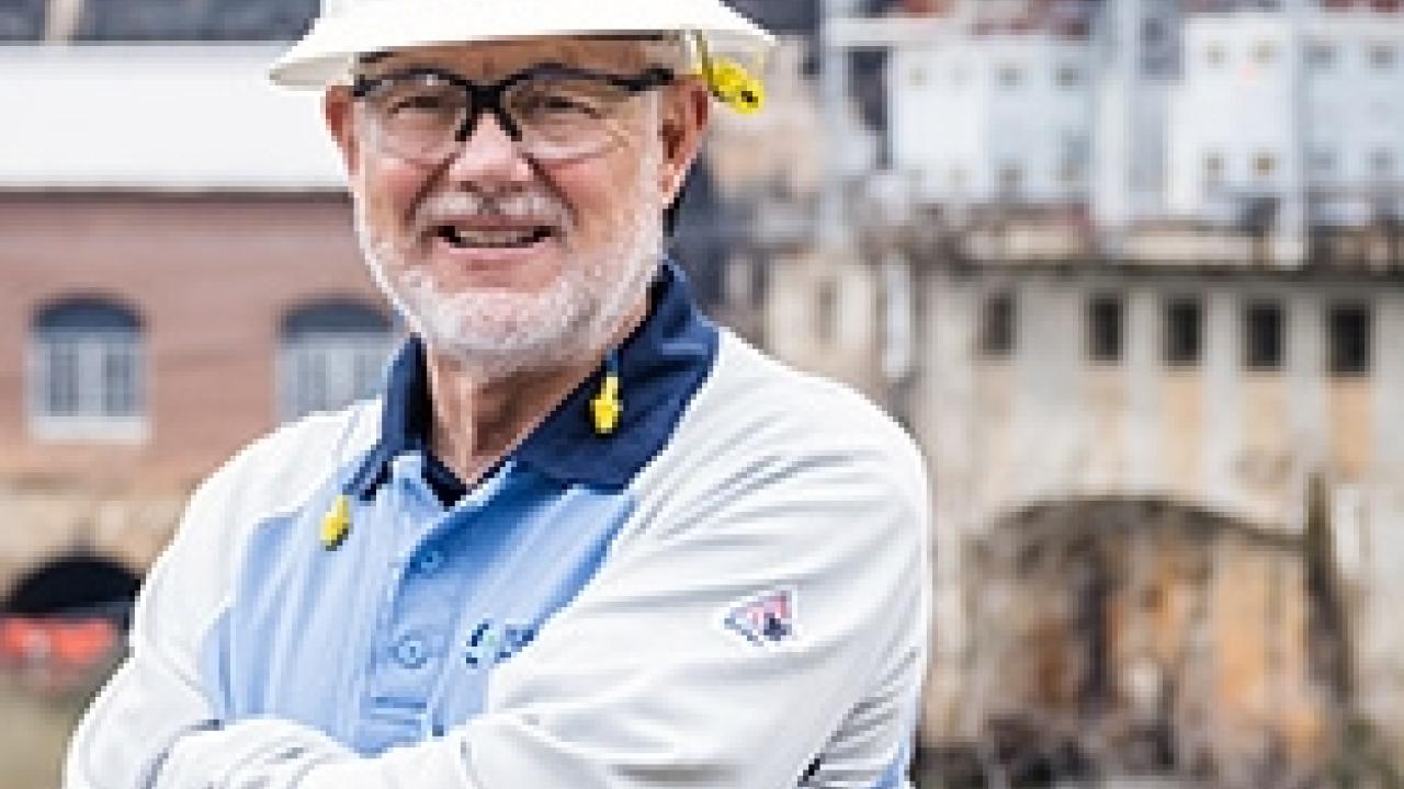 A smiling person with arms crossed in a hard hat. A hydroelectric plant behind them.