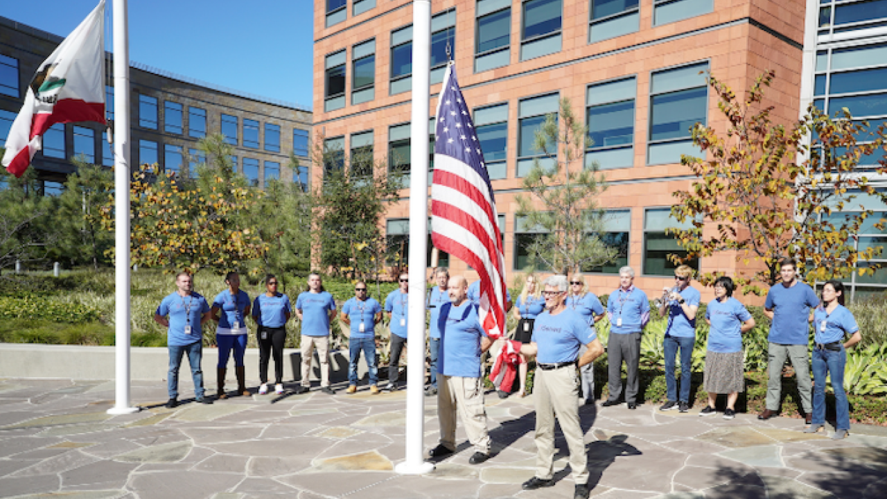 Veterans Day 2021 flag-raising ceremony at Illumina headquarters. Two males are raising the flag and a group of Illumina veterans are in a semi-circle around the back of the flag pole. The crowd is made up of 12 men and women.