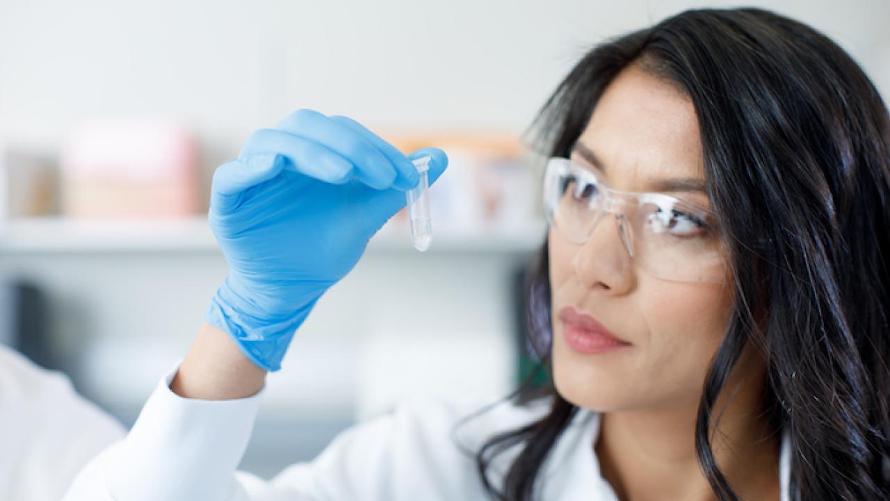 Illumina female scientist in white gown, gloves and protective eyewear looks at a small sample in a test tube.