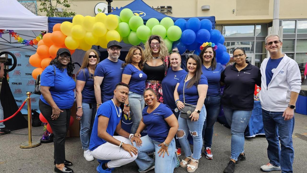 Colleagues in matching shirts in front of a rainbow balloon arch. All wearing rainbow lanyards.