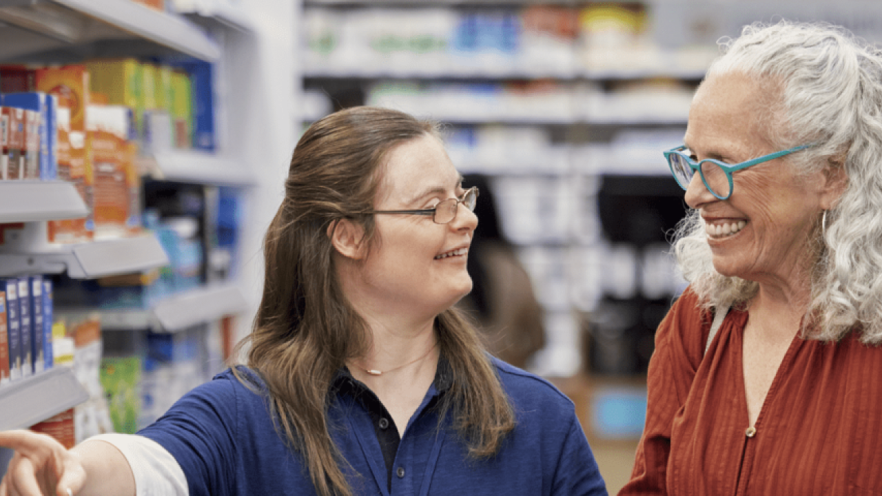 Two people smiling at each other in a pharmacy aisle, one points to an item on a shelf.