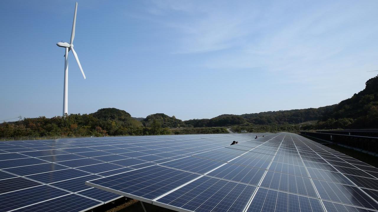A long row of solar panels. A wind turbine in the distance.