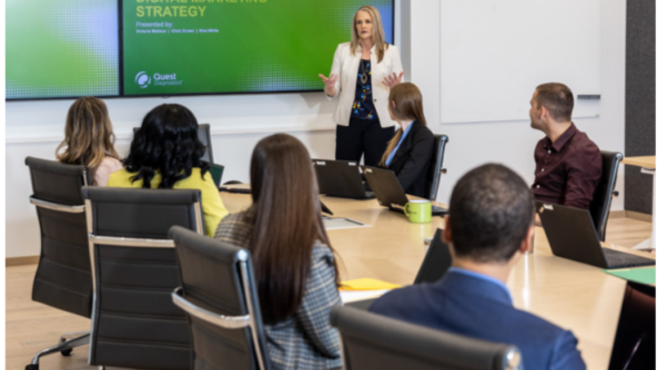 A person presenting to a room of others seated at a long table. A screen behind them.