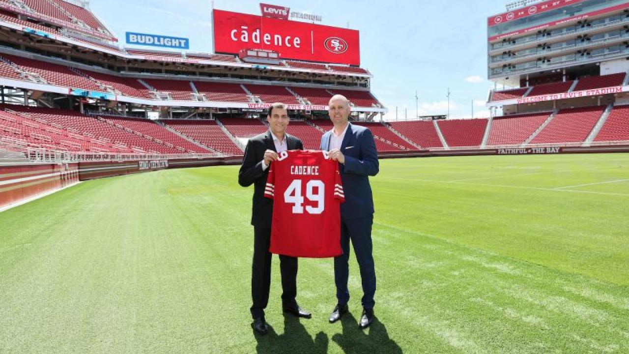 Two people holding a red jersey with 49 on it, standing on a football field. Large "cadence" billboard at the top of the stands.