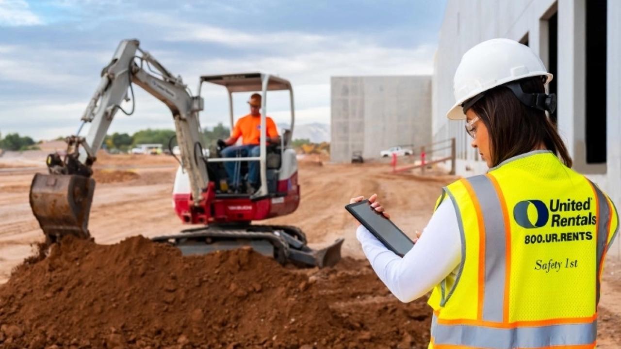 A person looking at a tablet. Another in the distance using a large digging machine.