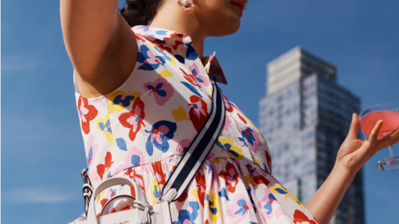 A person in a colorful kate spade dress, sunglasses, and cross-body handbag. A tall building behind them. 