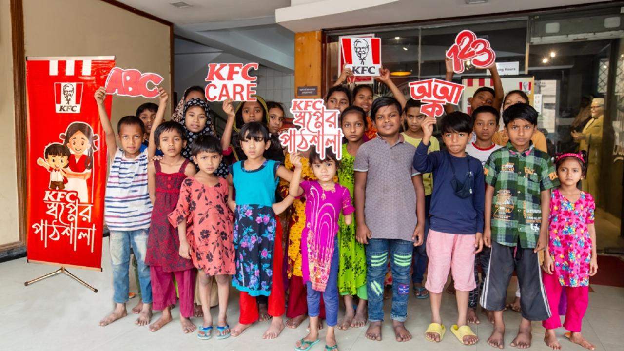 A group of standing children, holding signs of the KFC logo, KFC Care, ABC, 123, and those in another language.
