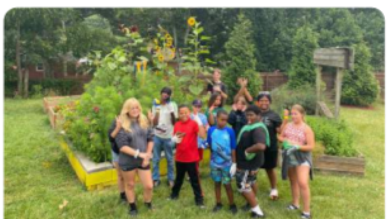 Kids and adults, some with raised hands, posed outside in front of a garden.