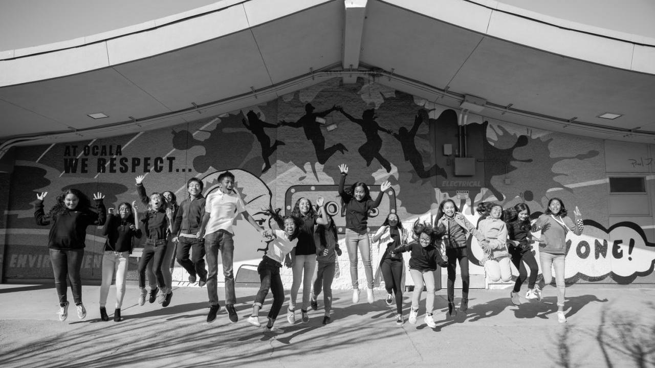 Black and white photo of a group of kids in a line jumping. A covered stage behind them.