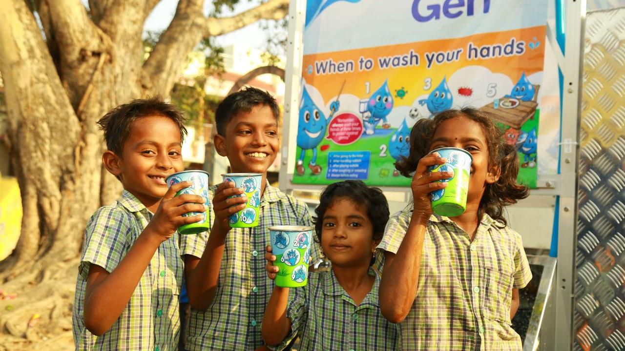 A group of smiling children holding out cups. A sign "Gen. When to wash your hands" behind them.