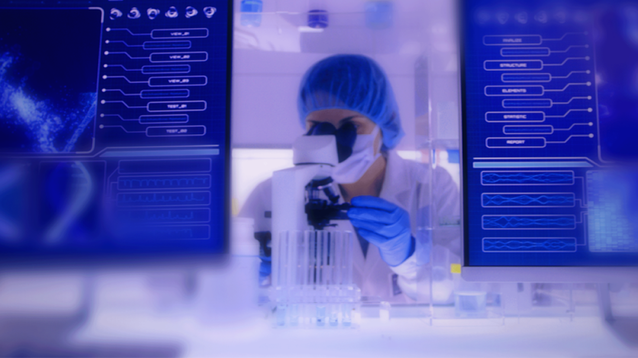 A person in medical protective wear looking in a microscope. Two computer screens to the sides.