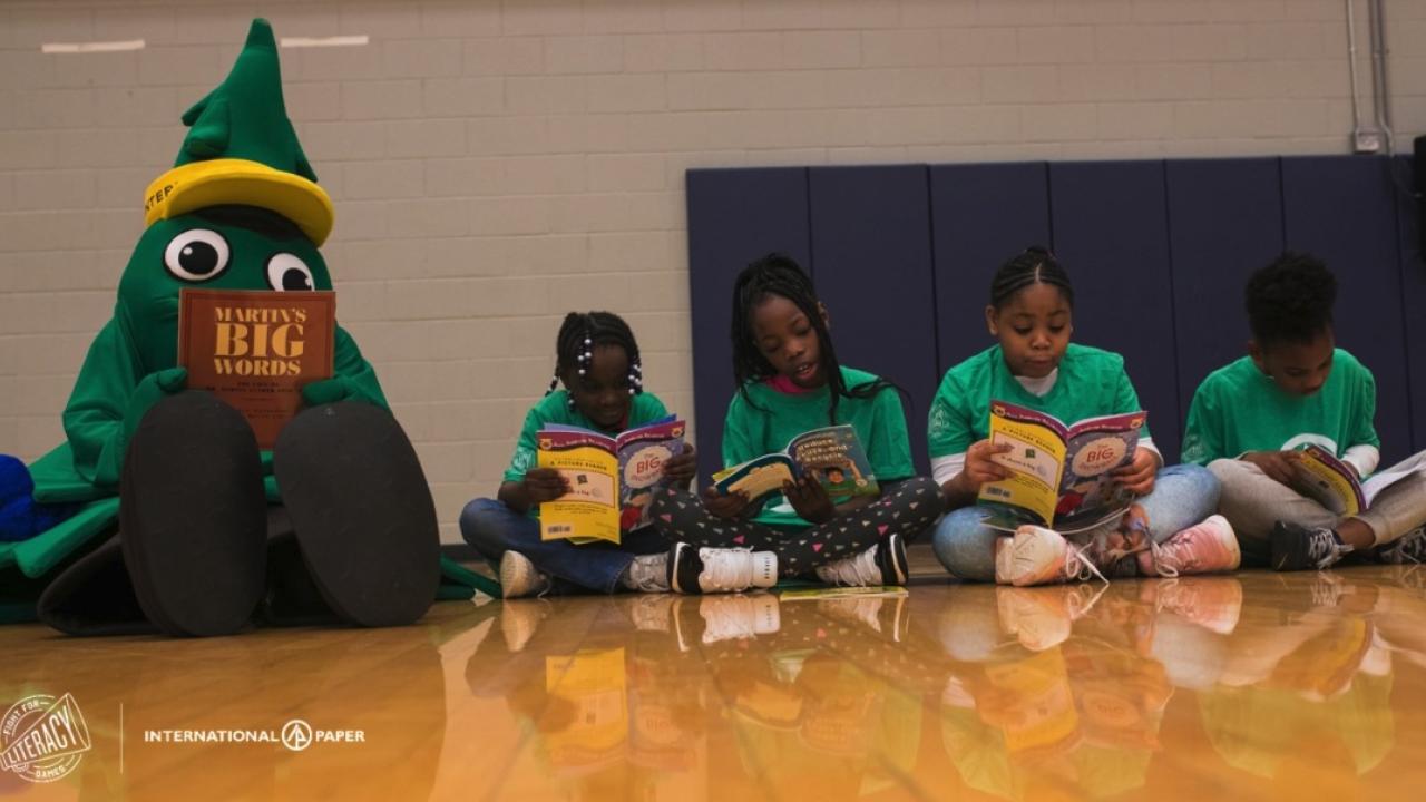 Children sitting in a line on a gym floor, a tree mascot next to them, all reading books.