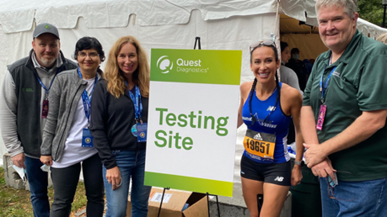 a group of five people, one in running apparel, by a sign for Quest "Testing Site" and a white tent behind them