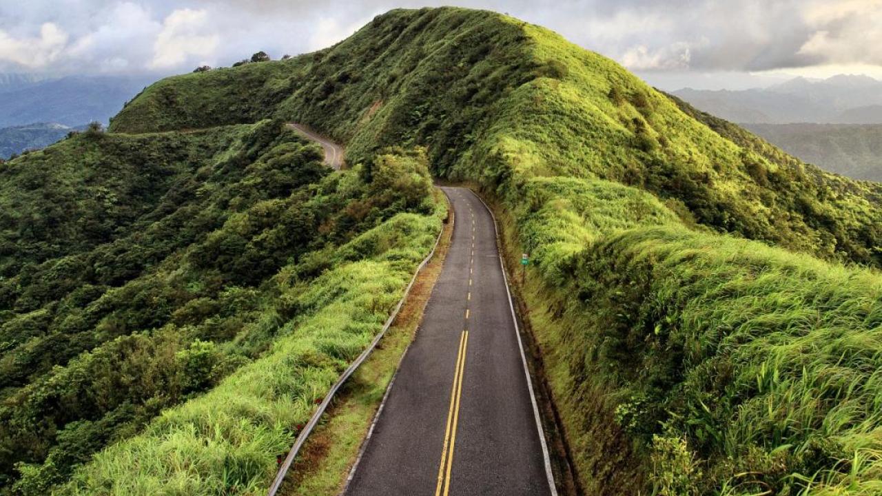 A two lane paved road leading through a greenery-covered peak. Cloudy sky in the background.