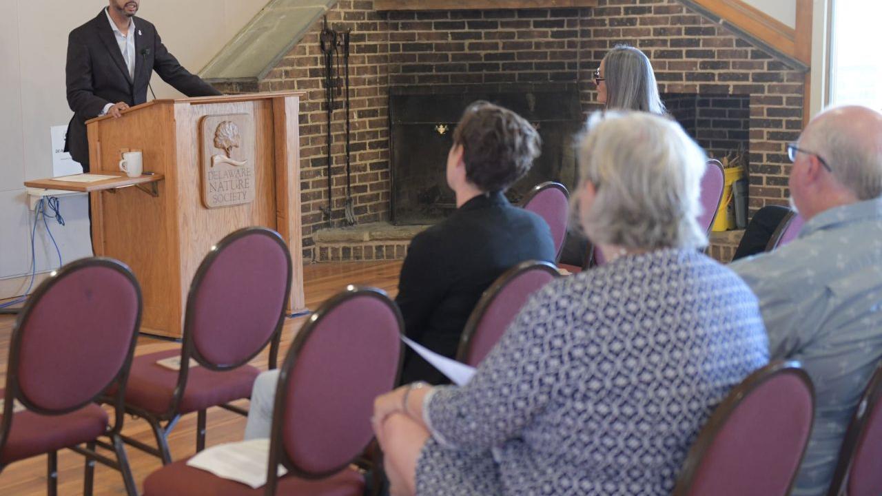 A person speaking at a podium in a room of others seated.