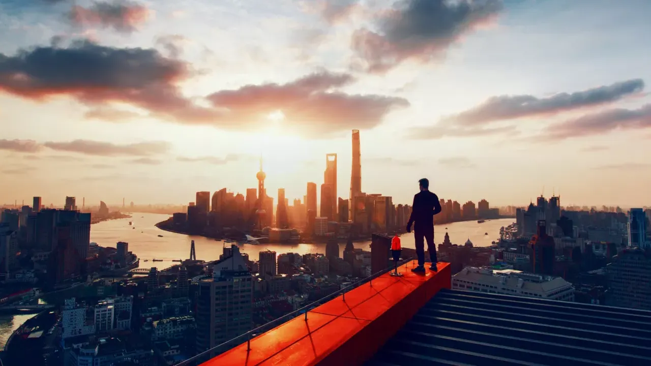 A person standing on a construction beam on the top of a very tall building, overlooking a city skyline and river.