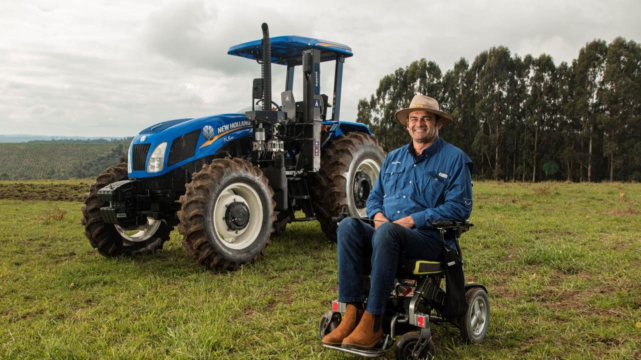 A person in a wheelchair in front of a New Holland tractor in a field.