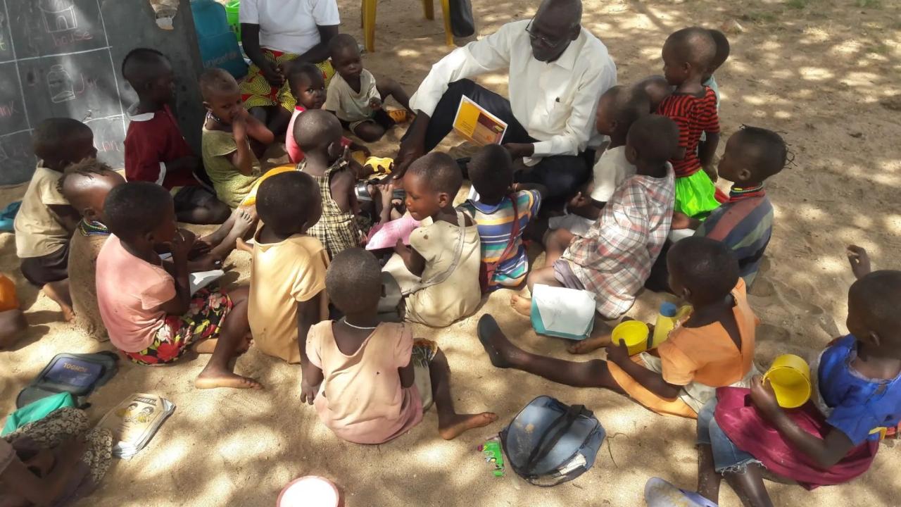 A group of children sitting on sandy ground, an adult showing them a picture