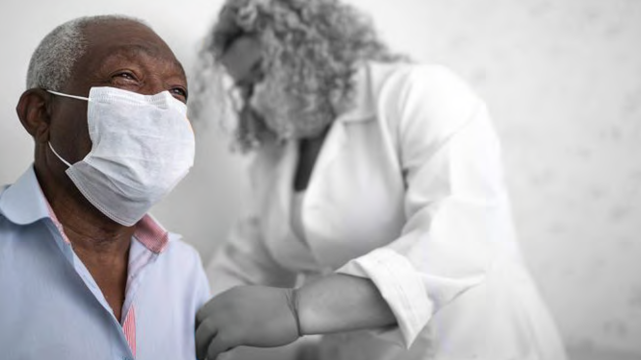 a medical professional looks at the left arm of another person. Both are wearing protective masks