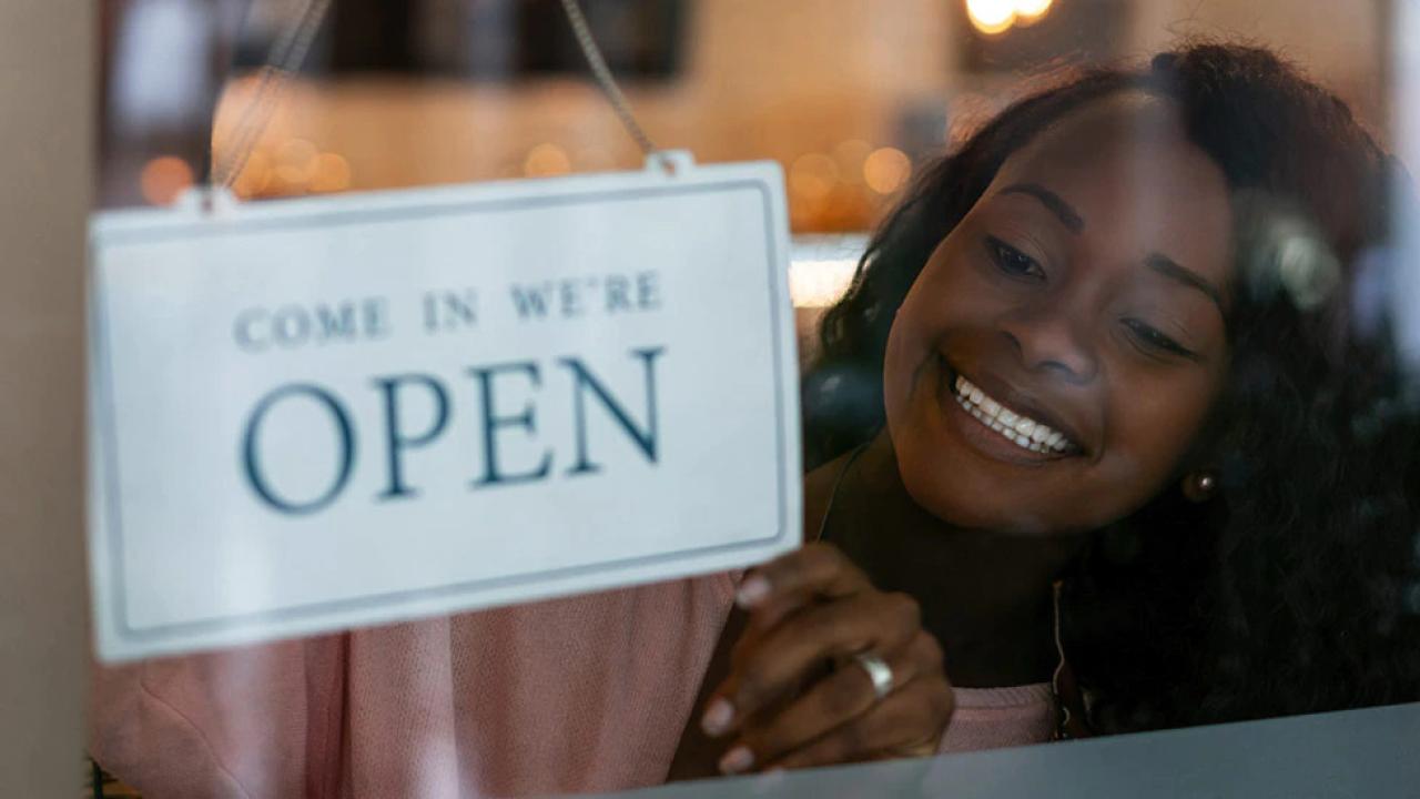 A smiling person flipping a sign to say "Come in we're OPEN" 