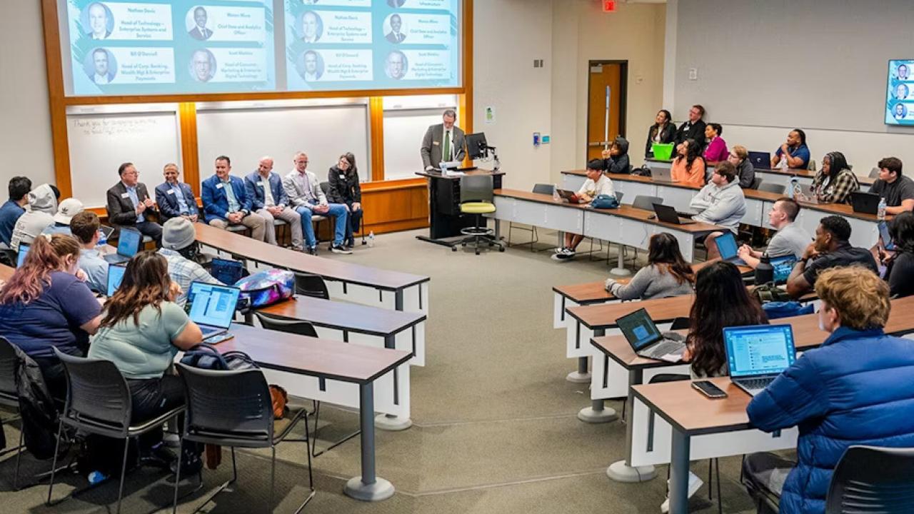 A speaker at a podium in a room of seated others. A digital display behind them with profiles of the panelists.