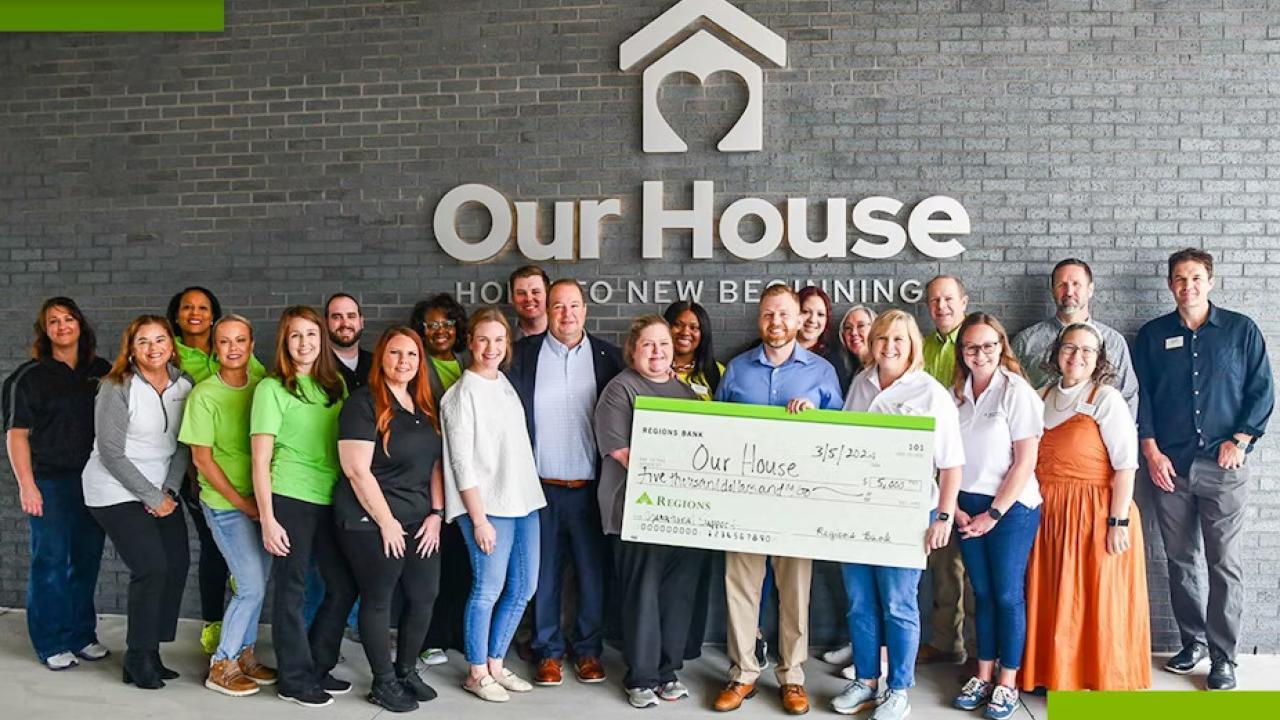 A greoup of people posed in front of the side of a building "Our House" sign above them. Some holding a large check made to "Our House".