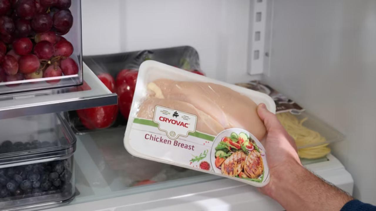 A hand holding a package of "Chicken Breast" in a refrigerator.