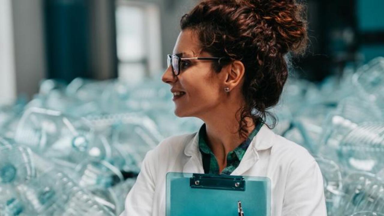 A person in a lab coat with a clip board in front of a large pile of clear plastic containers.