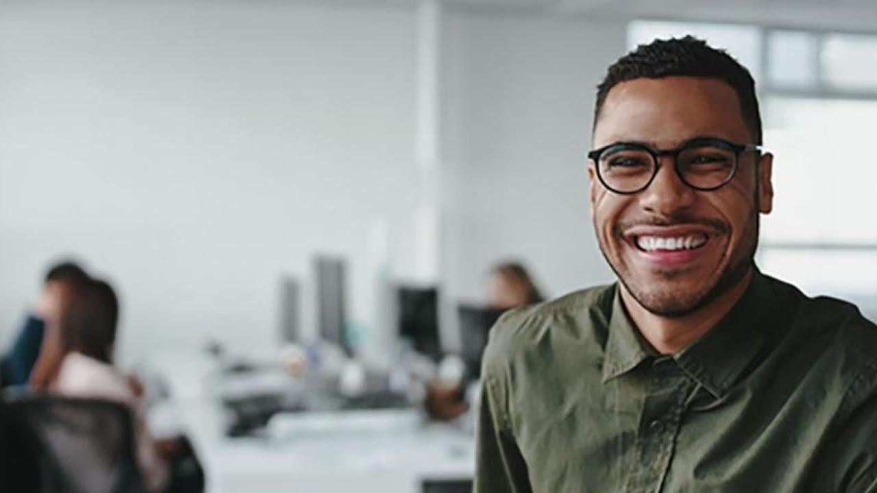 A smiling person looking at the camera, a small group of people at a shared desk behind them, in an office setting