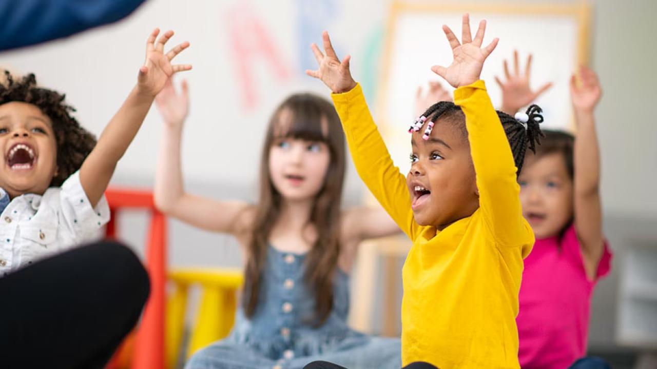 Children seated in a classroom setting, all with raised hands.