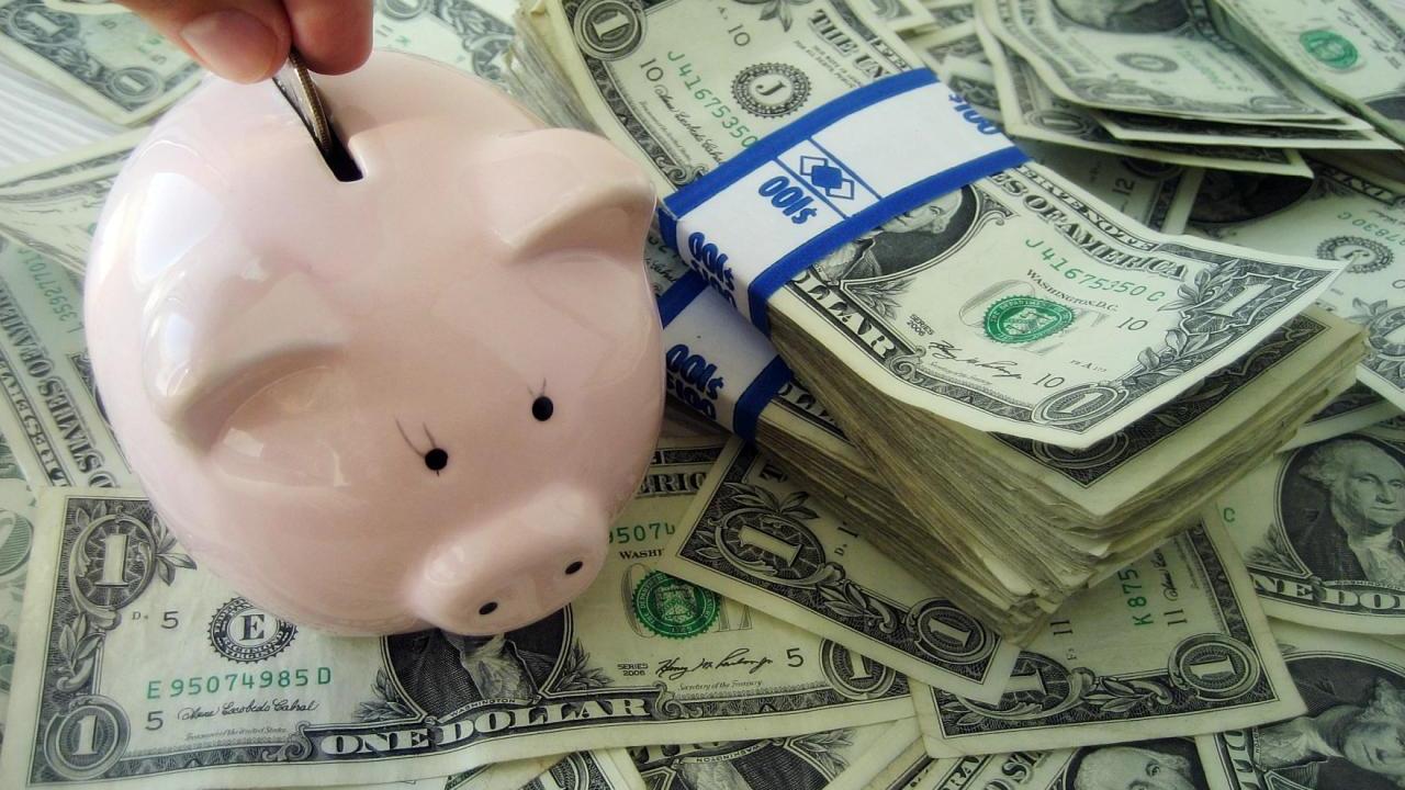A hand putting a coin in a small piggy bank atop stacks of dollar bills.